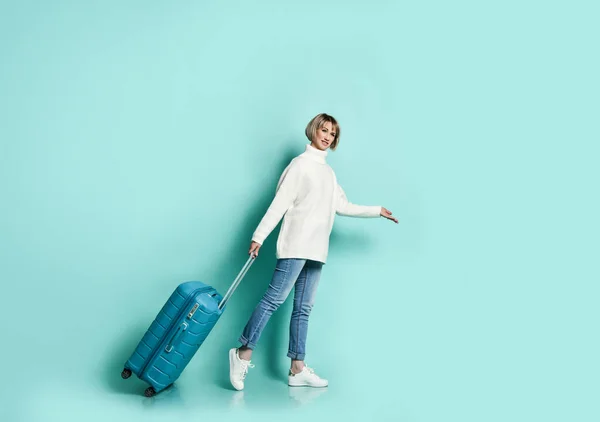 Blonde girl in white sweater, jeans and sneakers. She is smiling posing with suitcase luggage standing sideways on blue — Stock Photo, Image