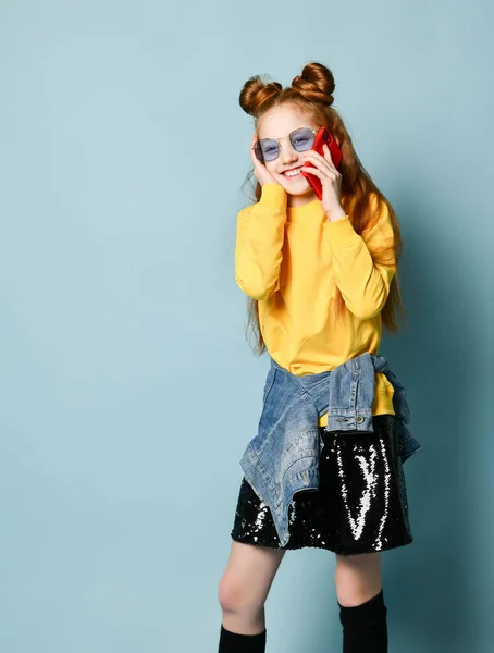 Teen red-haired girl in blue smoked glasses, yellow sweatshirt and glossy black skirt is talking by phone giggling — Stock Photo, Image