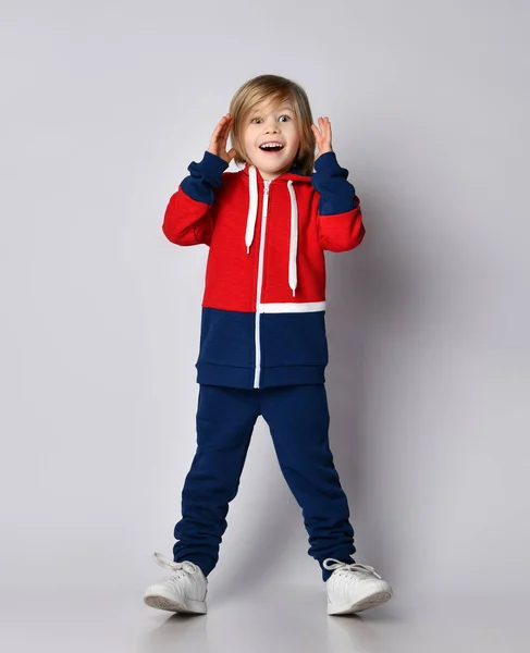 Happy excited surprised blond kid boy in red and blue sportwear with wide opened eyes holds his hand at her head — Stock Photo, Image