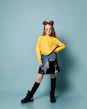 Havalı kızıl saçlı genç kız mavi füme gözlük, sarı kazak, parlak siyah etek ve vahşi ayakkabılar içinde poz veriyor.