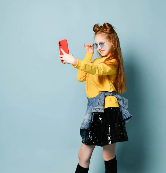 Mavi füme gözlüklü, sarı kazaklı ve parlak siyah etekli kızıl saçlı genç kız akıllı telefonuyla selfie çekiyor. — Stok fotoğraf