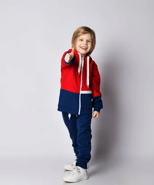 Happy smiling blond kid boy in blue and red sportswear hoodie and pants shows thumb up sign gesture — Stock Photo, Image