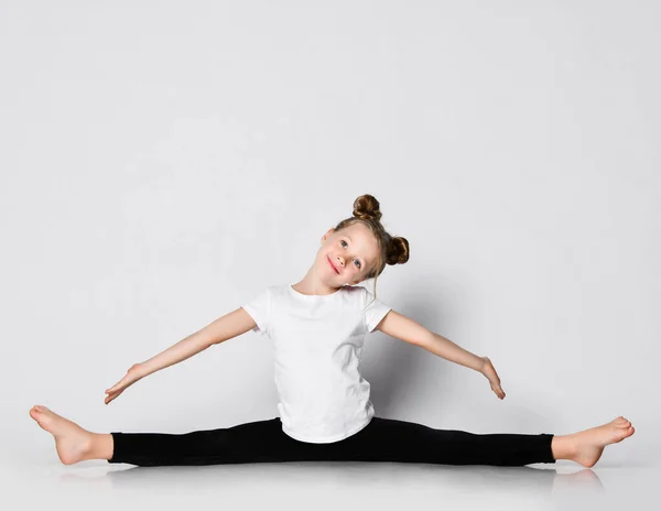 Sonriente niña hace ejercicios de gimnasia en casa se divide con los brazos extendidos y la cabeza inclinada a un lado —  Fotos de Stock