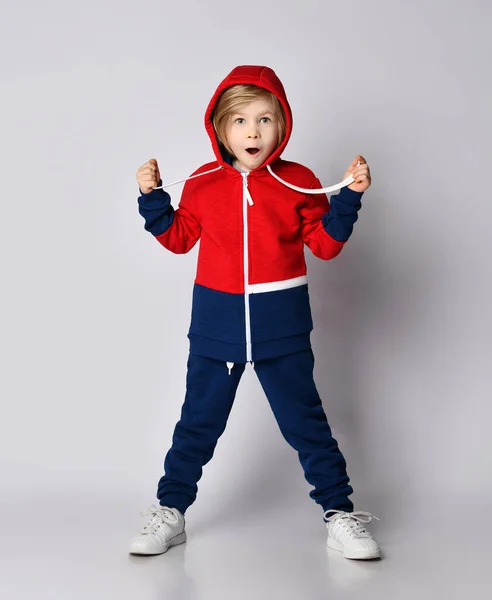 Surpreendido garoto loiro animado em azul e vermelho terno esportivo posa com capuz na cabeça — Fotografia de Stock