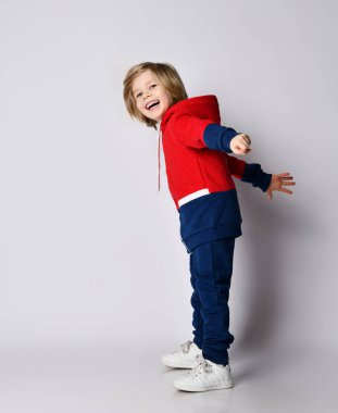 Kırmızı ve mavi spor takım elbiseli neşeli sarışın bir çocuğun tam boy portresi. Kolları açık bir şekilde yanımızda duruyor.