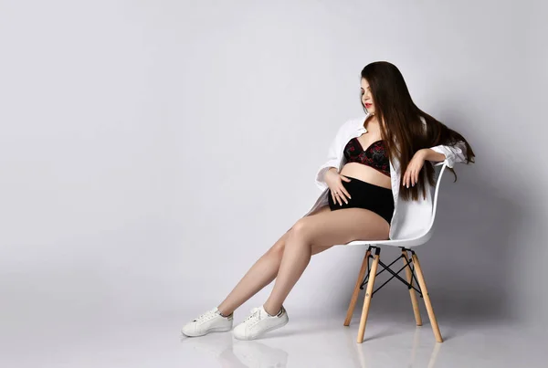 Fiatal szép karcsú terhes nő hosszú haj fehér ing ül a modern széken tartsa hasa fehér — Stock Fotó