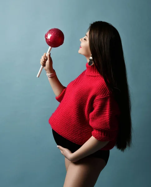 Gyönyörű terhes lány piros pulóverben egy hatalmas nyalókát tart pasztell kéken pózolva. — Stock Fotó