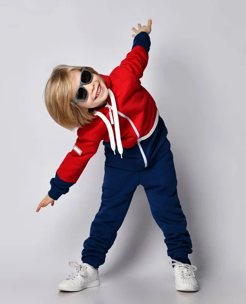 Sonriente chico rubio juguetón activo en sudadera con capucha azul y roja, pantalones y gafas de sol hace ejercicios de gimnasio —  Fotos de Stock