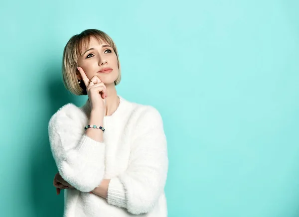 Thoughtful blonde woman in white sweater holds hand at her head looks up in corner at free copy space — Stock Photo, Image