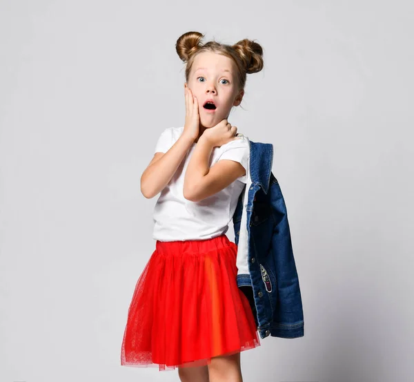 Geschokt blond kind meisje met grappige broodjes in rode rok en wit t-shirt poseert zijwaarts naar ons houden hand op haar wang — Stockfoto