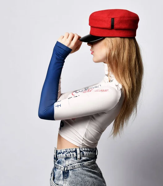 Jovem bela mulher loira magro em manga comprida, jeans e chapéu vermelho de pé formas laterais e segurando cobertura para a cabeça — Fotografia de Stock
