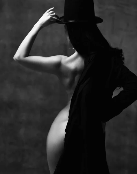 Νέα όμορφη μελαχρινή γυμνή γυναίκα με καπέλο στέκεται και κρατώντας μαύρα ρούχα στα χέρια πάνω από το σκοτάδι — Φωτογραφία Αρχείου