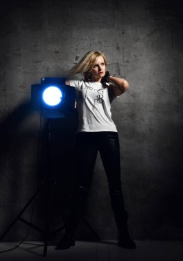 Şık giyimli ve siyah çizmeli güzel sarışın kadın aydınlık armatürünün yanında duruyor.