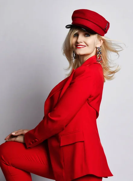 Portrét mladé krásné blond usměvavé ženy ve stylovém elegantním tmavočerveném oficiálním obleku, červené čepici a masivních výdělcích — Stock fotografie