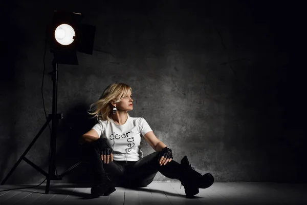 Молодая красивая блондинка в стильной повседневной одежде и сапогах, сидящая на полу возле светильника — стоковое фото