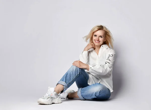 Young beautiful positive blond woman in stylish casual clothing and white sneakers sitting on floor and smiling — Stock Photo, Image