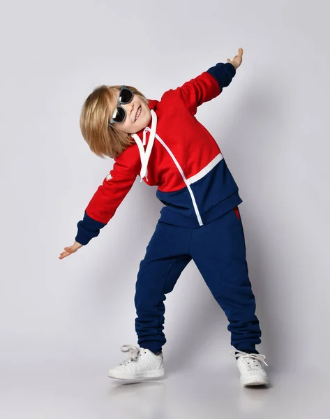 Alegre activo juguetón rubio niño en azul y rojo sudadera con capucha, pantalones y gafas de sol está jugando piloto de avión —  Fotos de Stock