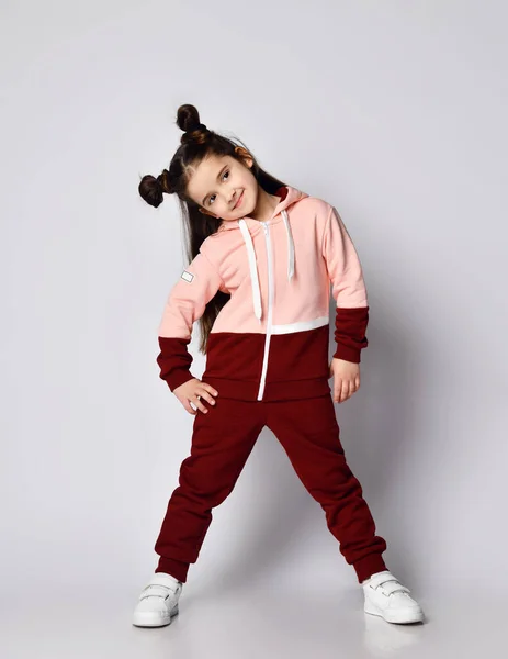 Sly smiling brunette kid girl with buns in modern stylish pink brown sport suit is posing with her head bowed aside