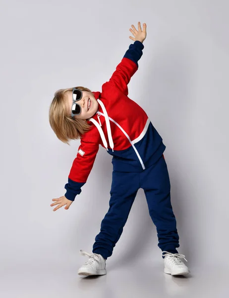 Happy smiling active blond kid boy in blue and red hoodie, pants and sunglasses does gym exercises — Stock Photo, Image
