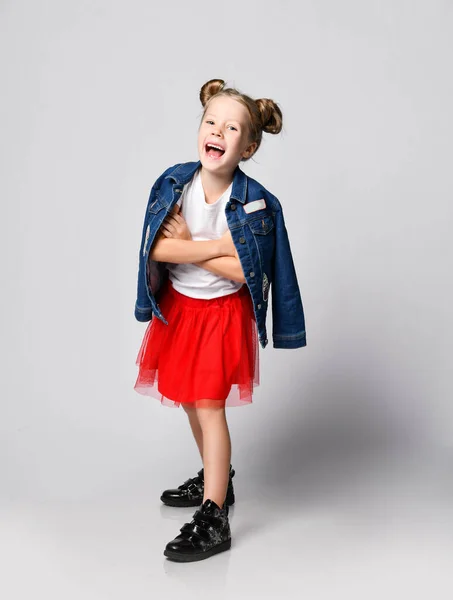 Blonde kid girl with funny buns in red skirt, white t-shirt and denim jacket over shoulder poses having fun — Stock Photo, Image