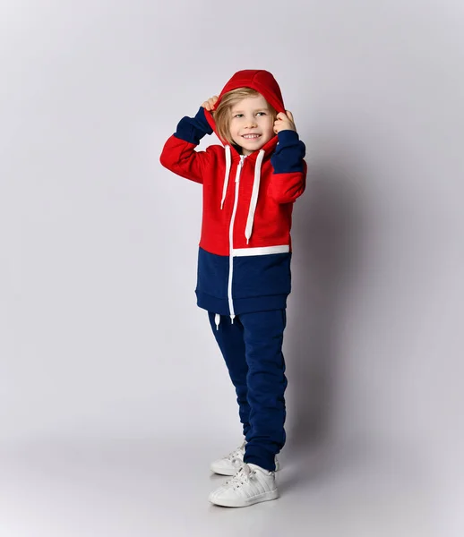 Smiling blond kid boy in blue and red sport suit puts on a cowl of his hoodie on his head — Stock Photo, Image