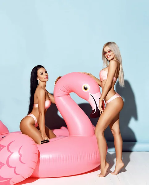 Sommerstimmung. Zwei aufgeregte junge schlanke Mädchen sitzen auf einem aufblasbaren rosa Flamingo-Spielzeug-Float und haben Spaß im gelben Bikini — Stockfoto