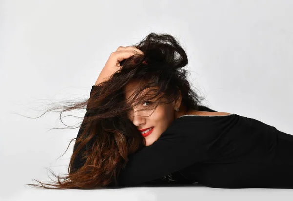 Flirting brunette woman in black dress is lying posing on the floor covering head with hands hiding face in curly hair — Stock Photo, Image