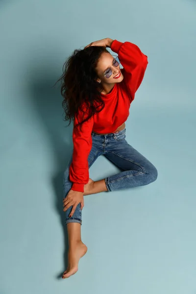 快乐的成年女性，一头卷曲的黑发，戴着烟熏的蓝色眼镜、红色毛衣和蓝色牛仔裤，坐在地板上 — 图库照片