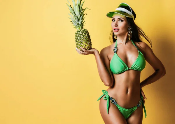 Jeune belle femme brune mince bronzée en bikini vert sexy avec décorations et casquette debout et tenant ananas frais — Photo