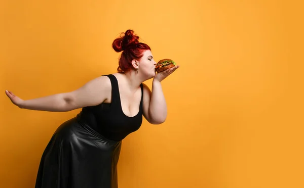 Positieve overgewicht dikke vrouw kust bijt hamburger cheeseburger sandwich met rundvlees ze houdt in de hand op geel — Stockfoto