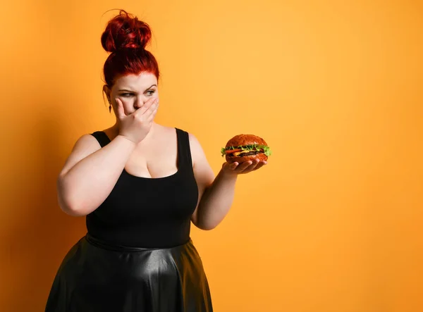 Overgewicht dikke vrouw kijkt met afschuw naar hamburger cheeseburger sandwich met rundvlees houdt ze in de hand op geel — Stockfoto