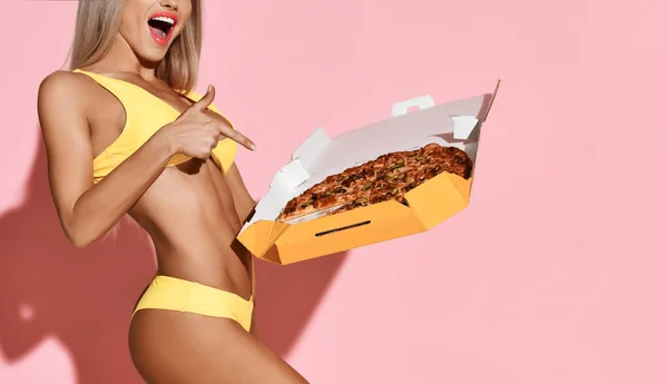 Jeune femme excitée blonde mince bronzée sexy en bikini de plage jaune sexy pointant vers la pizza fraîche entière dans la boîte qu'elle tient — Photo