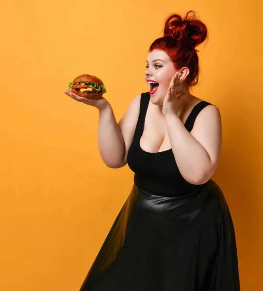 Happy overweight plus-size woman looks at burger cheeseburger sandwich with beef she holds on her open palm on yellow