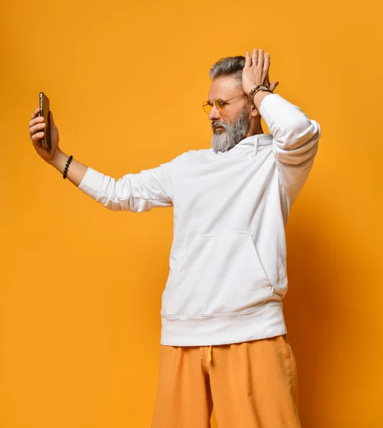 Stylish grey-haired hipster man in white hoodie and sunglasses standing making selfie on smartphone — Stock Photo, Image