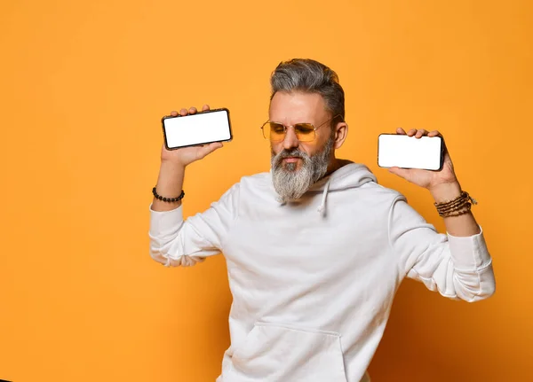 Elegante hombre hipster de pelo gris con capucha blanca y gafas de sol rojas de pie y sosteniendo dos teléfonos inteligentes en las manos —  Fotos de Stock