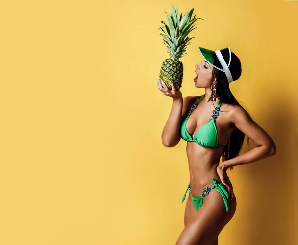 Jeune femme brune mince bronzée en bikini vert sexy avec des décorations et une visière va mordre un ananas frais — Photo