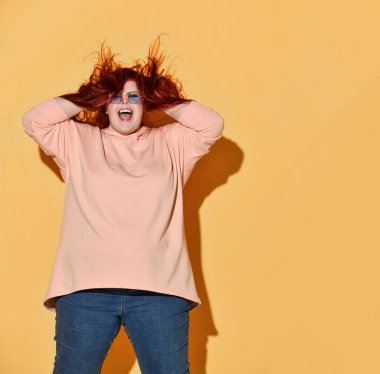 Genç, mutlu, kızıl saçlı, kilolu, sıradan giyinmiş, ayakta duran, bağıran ve saçlarıyla oynayan bir kadın.