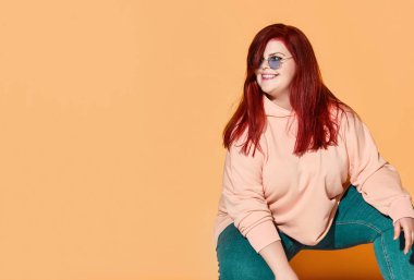 Gülümseyen kızıl saçlı, kilolu, günlük kıyafetler ve güneş gözlüğü takan mutlu hisseden ve yan bakan bir kadın.