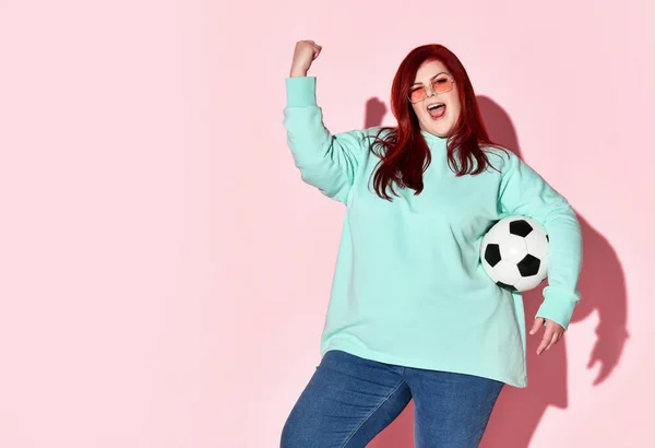 Junge schöne rothaarige übergewichtige Frau in Jeans, blauem Kapuzenpulli und Sonnenbrille, die mit Ball steht und Glück ausdrückt — Stockfoto