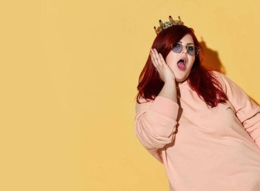 Genç, kızıl saçlı, kilolu, günlük kıyafetler giyen, güneş gözlüklü ve kafasında altın bir taç olan kadın şaşırmış hissediyor.