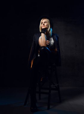 Gülümseyen sarışın kadın şık günlük tişörtlü siyah ceketli, deri eldivenli, pantolonlu ve şapkalı yüksek sandalyede oturuyor.