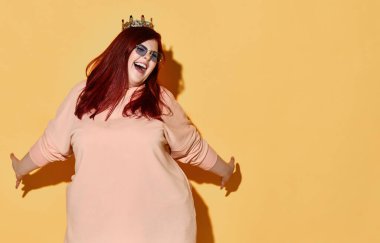 Neşeli, kızıl saçlı, kilolu, sıradan giysili, kafasında altın bir taç olan, kolları açık, neşeli bir kadın.