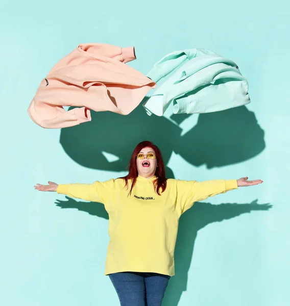 Joven mujer pelirroja feliz con sobrepeso en sudadera con capucha amarilla y vaqueros azules arroja hacia fuera la ropa azul y rosa — Foto de Stock