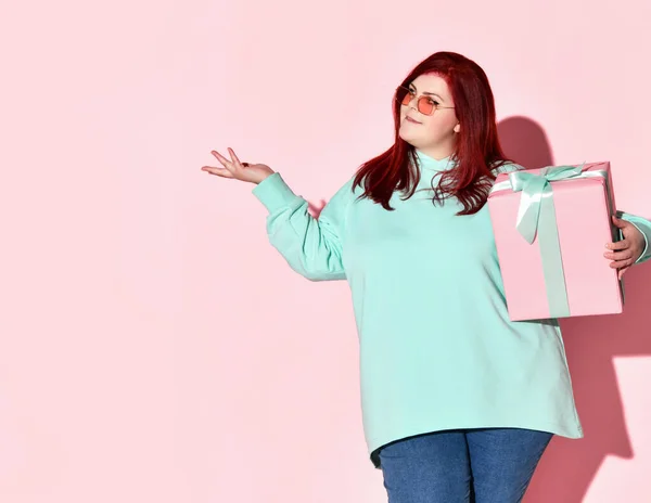 Mujer joven con sobrepeso en jeans, sudadera con capucha azul y gafas de sol de pie con caja de regalo sosteniendo la otra mano con la palma abierta — Foto de Stock