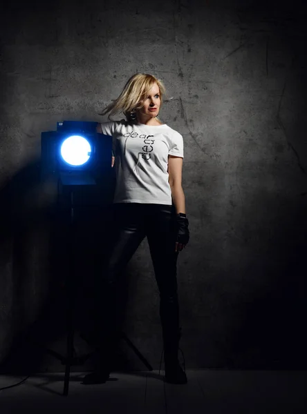 Mladá krásná blondýnka v neformálním tričku, kožených kalhotách, rukavicích bez prstů a botách stojící ve světle reflektorů — Stock fotografie