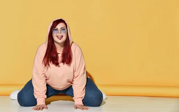 Fiatal játékos túlsúlyos nő elegáns kapucnis kapucnis csuklyával a fején, kék farmer és napszemüveg pózol a padlón — Stock Fotó