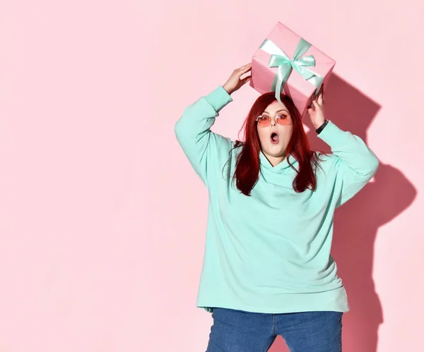 Happy surprised overweight woman in blue jeans, hoodie and sunglasses holding gift box on her head