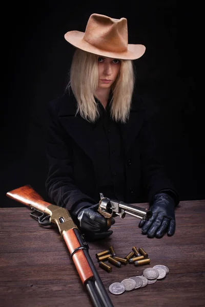 狂野的西部金发姑娘 拿着枪坐在桌子旁 枪弹和银币 背景是黑色的 — 图库照片