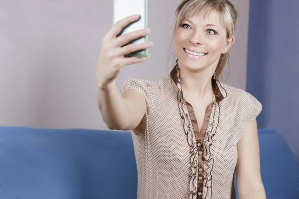 Cute sexy blond girl sits on sofa. She looks at her smartphone and makes a selfie. — Stock Photo, Image