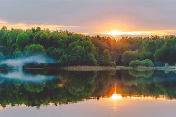 Una puesta de sol tranquila en la orilla del lago, donde el bosque se refleja en aguas tranquilas con neblina y niebla — Foto de Stock